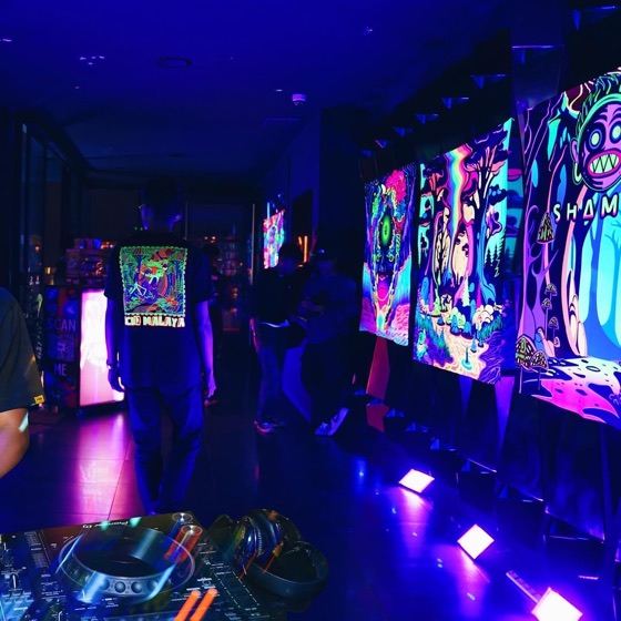 Acid Malaya tee glowing under UV light beside a wall of blacklight art